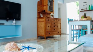 A dining area with a glass table, a wooden cabinet, a TV mounted on the wall, and a blue chair. - Klima Paradise (Samos)