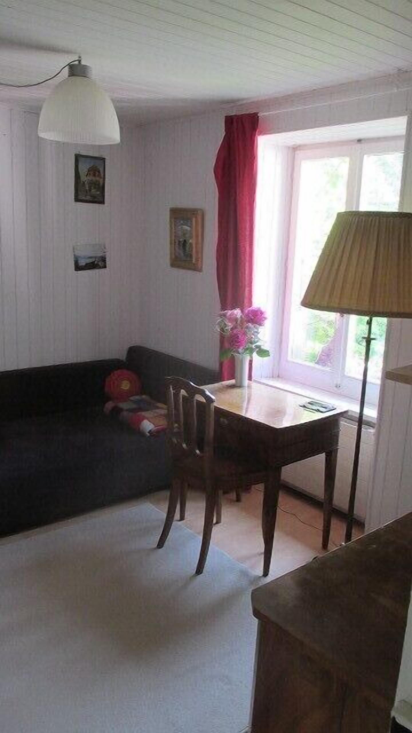 Une petite pièce agréable et chaleureuse, avec un canapé, une table et une lampe. Des tableaux ornent le mur et une fenêtre est agrémentée de rideaux.