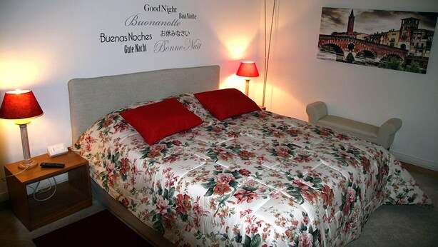 A neatly made bed with floral bedding and red pillows, a bedside table with a lamp, and a wall with multilingual "Good Night" message. - B&B al 19 (Verona)