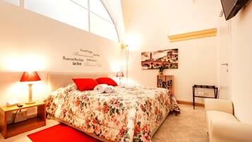 Une chambre avec un grand lit, des oreillers rouges, un tapis rouge, une table de chevet avec une lampe et un mur orné d’un tableau représentant un paysage urbain.