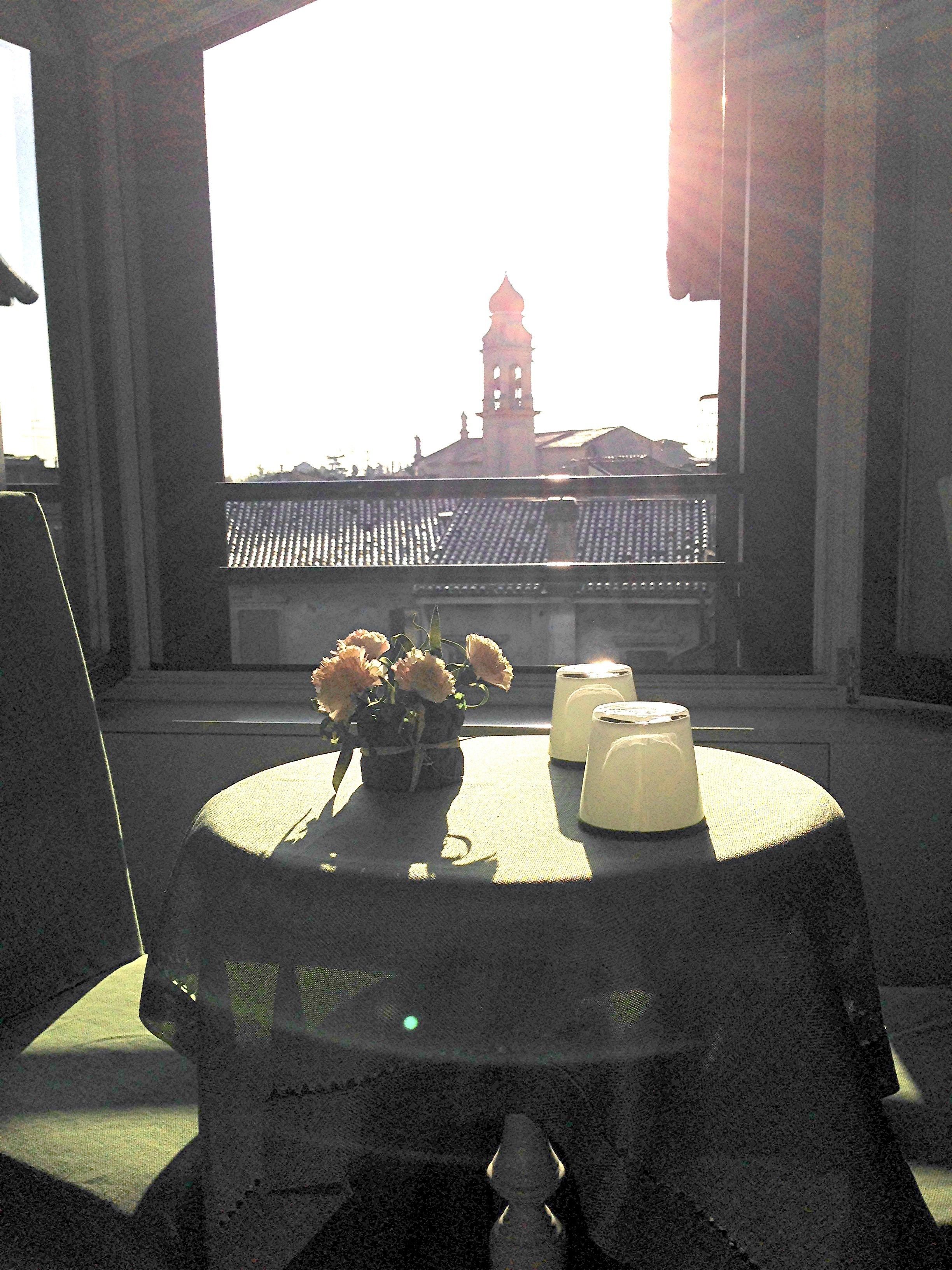 A table with a vase of flowers and candles, a window with a view of a building with a clock tower, and a balcony with a tiled roof.