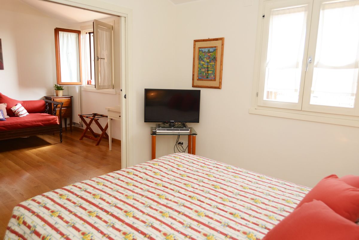 A bedroom with a bed, a television on a stand, a window, and a door leading to another room.