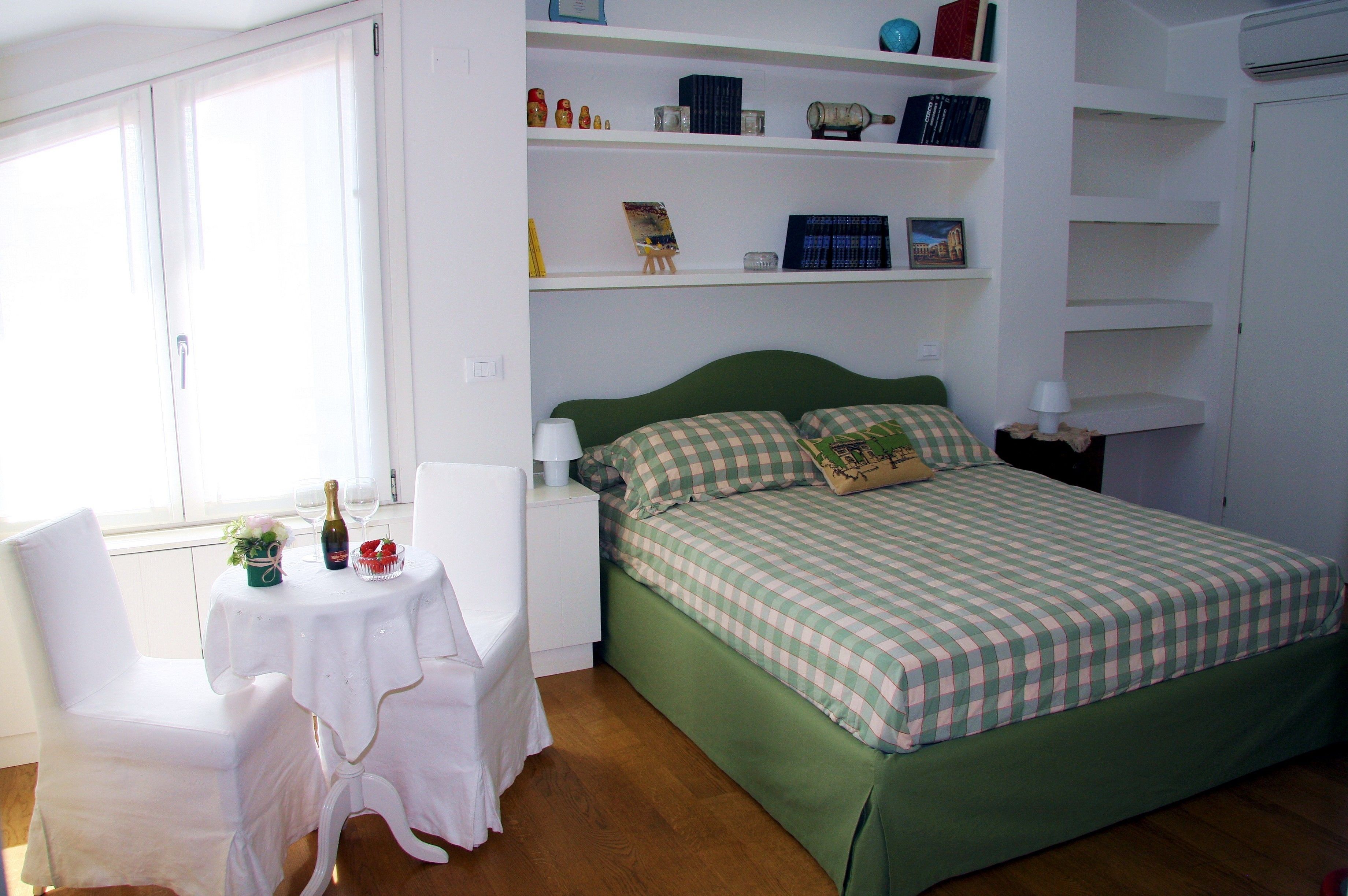 Een slaapkamer met een groen bed, witte muren en een raam. Er staat een klein tafeltje met een fles en glazen, en een stoel.
