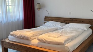 A wooden bed with white bedding in a room with a window and a wall-mounted lamp. - Haus Ares (Zermatt)