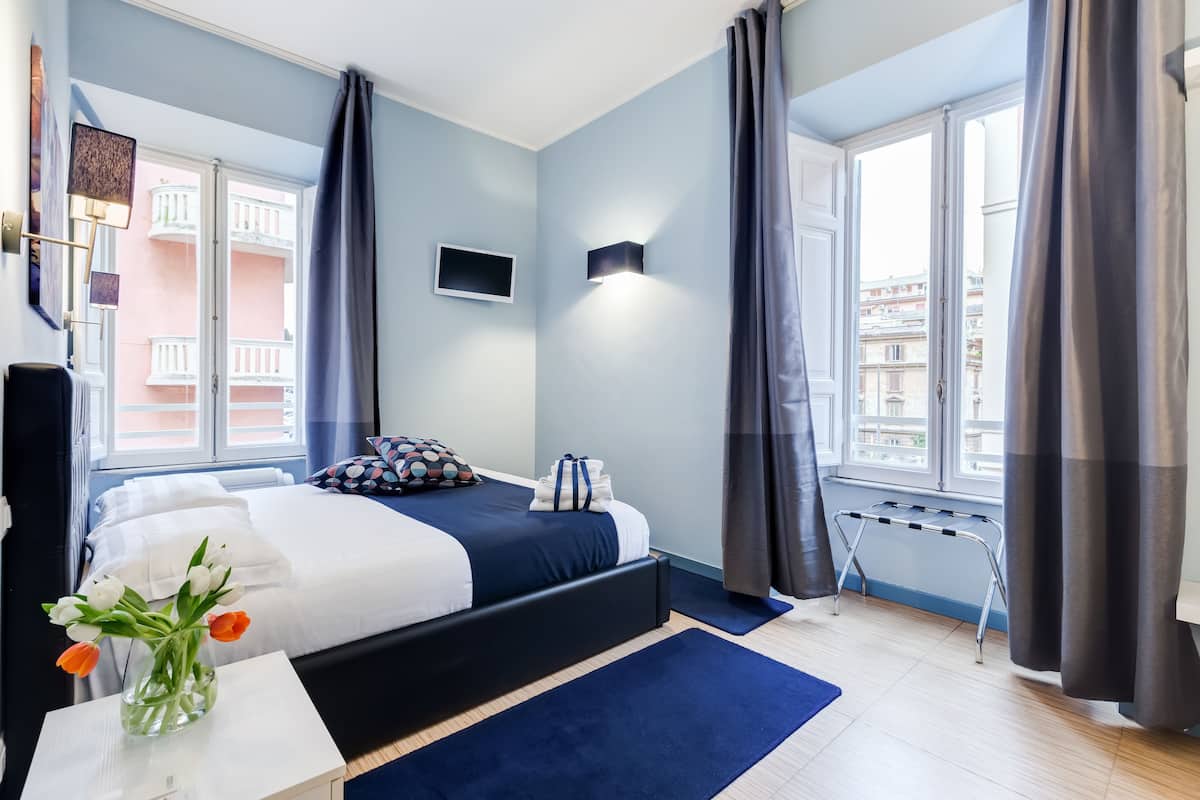 Habitación de hotel moderna con cama, una mesita con un florero, televisor y ventana con cortinas.