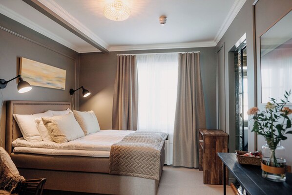 A bedroom with a bed, bedside lamps, a wooden bench, a vase with flowers, and a painting on the wall.