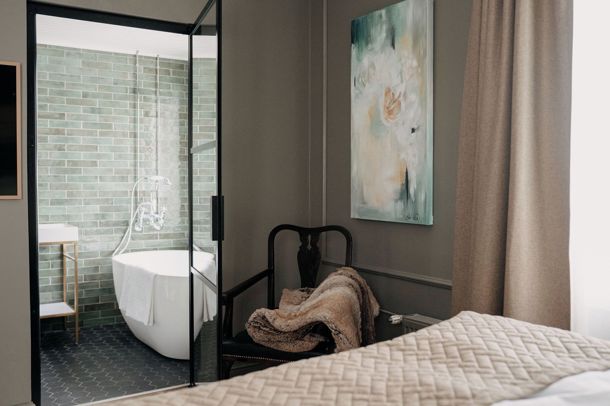 Ett hotellrum med en glasdörr som leder till ett badrum med ett vitt badkar och en dusch. Det finns en säng med ett vadderat täcke och en stol med en filt.