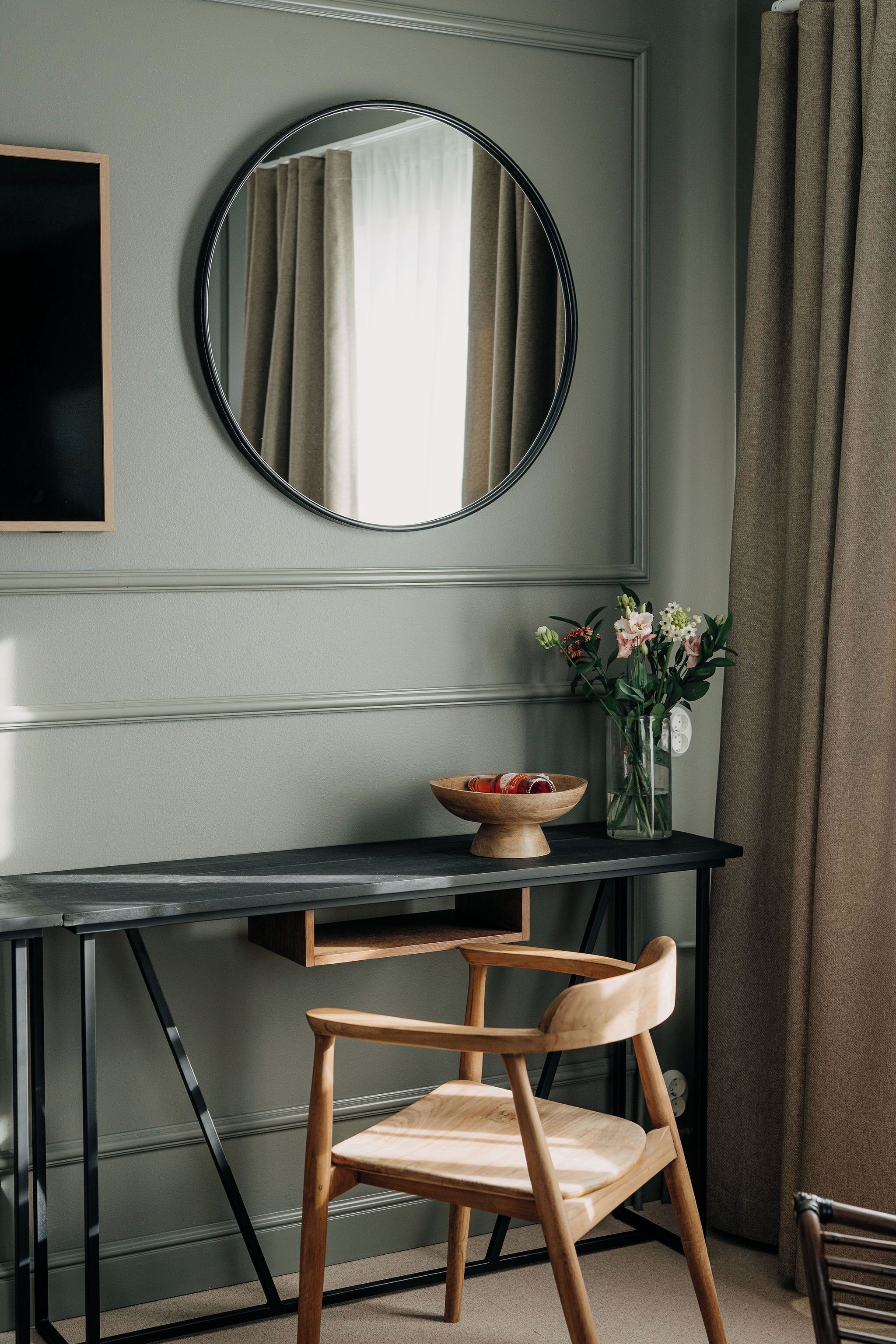 Ein Zimmer mit einem runden Spiegel, einem Holzstuhl, einem kleinen Tisch mit Schale und Blumen sowie einem dunklen Schreibtisch an einer hellen Wand.