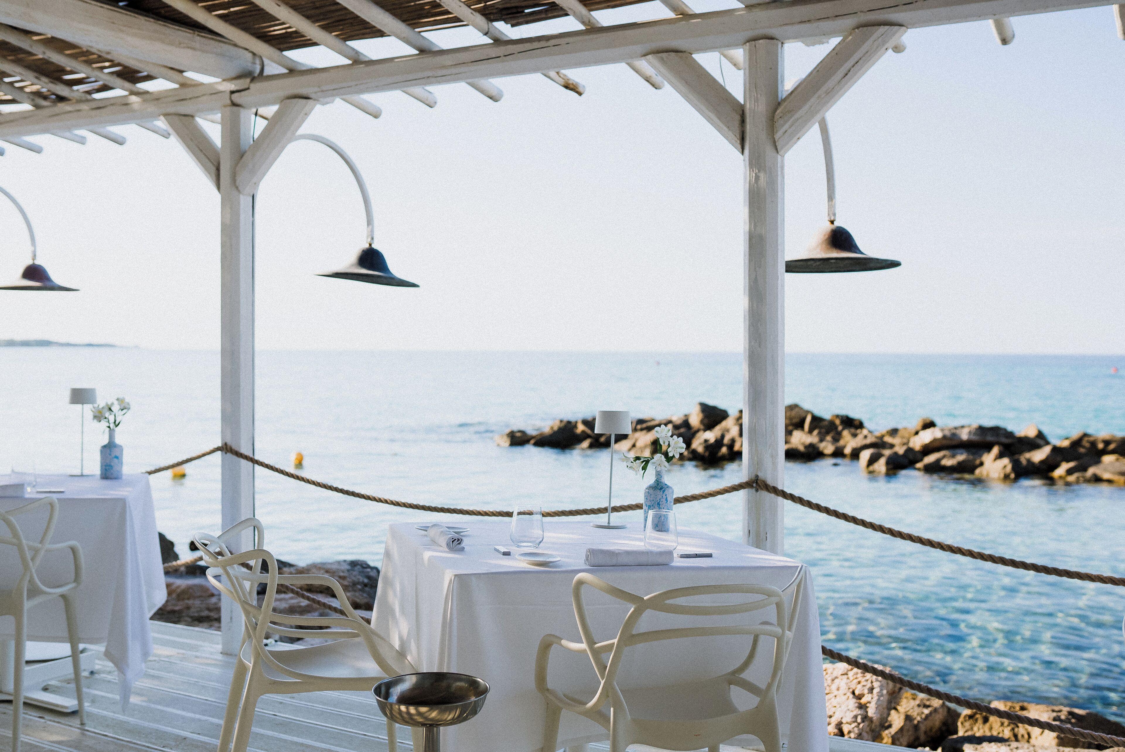 A seaside restaurant with white tables and chairs, a rope barrier, and a view of the sea and rocks.