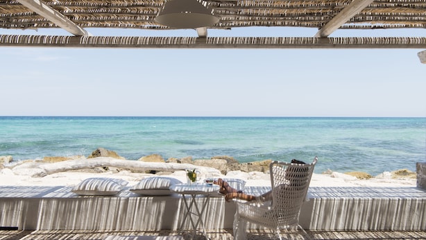 A beachside seating area with a view of the ocean, a table with a drink, and a straw hat on the ground. - La Peschiera (Monopoli)