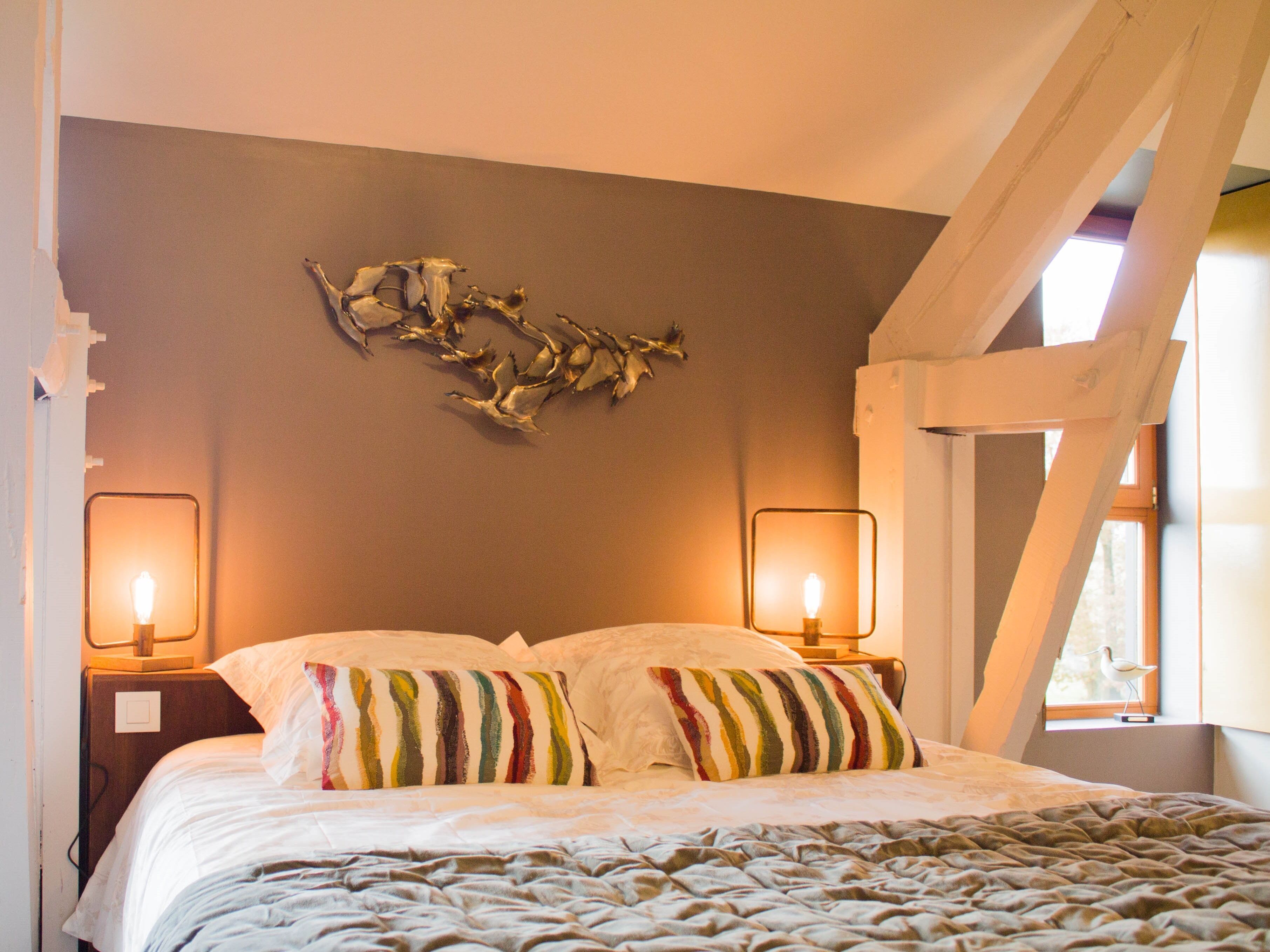 A bedroom with a bed, two bedside lamps, a wall-mounted abstract art piece, and a window with a view of trees.