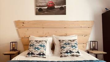 A bedroom with a wooden headboard, two patterned pillows, a blue blanket, and a painting of an airplane on the wall.