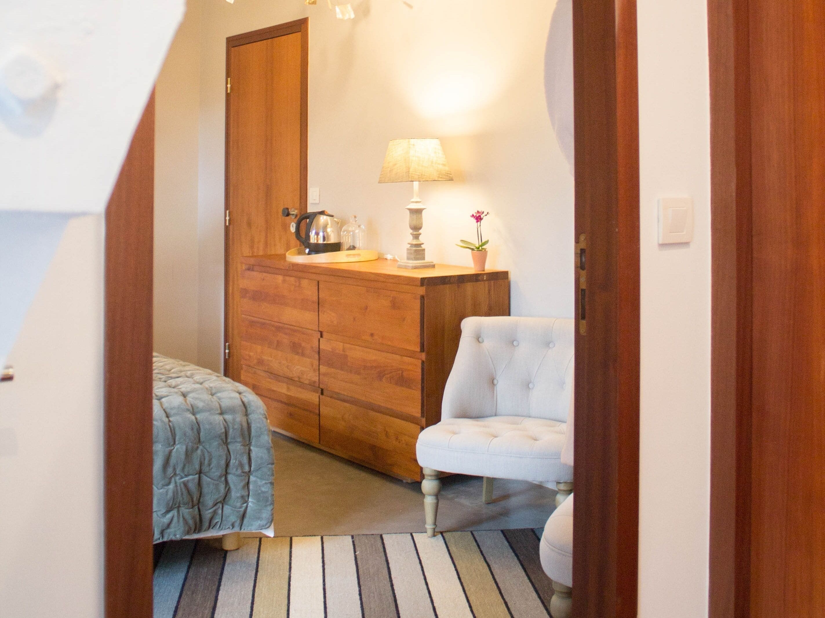 A narrow hallway with a wooden door, a bed with a blue quilt, a white upholstered chair, a wooden dresser with a lamp and a potted plant.