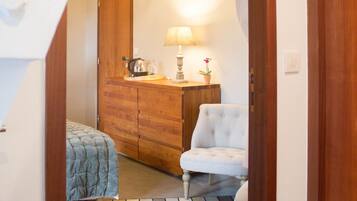 A narrow hallway with a wooden door, a bed with a blue quilt, a white upholstered chair, a wooden dresser with a lamp and a potted plant.
