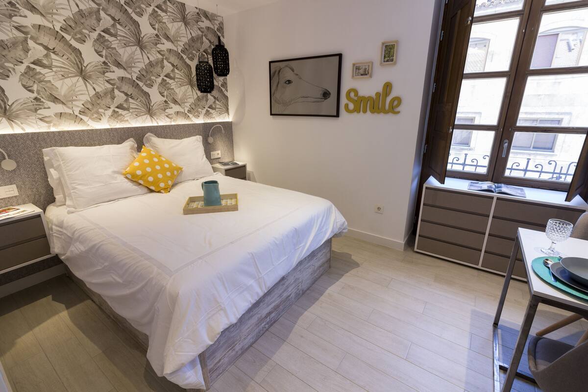 Une chambre à coucher bien aménagée, avec un lit, des tables de chevet, une fenêtre donnant sur un beau paysage et un mur orné d’un motif de feuilles et d’un tableau encadré.