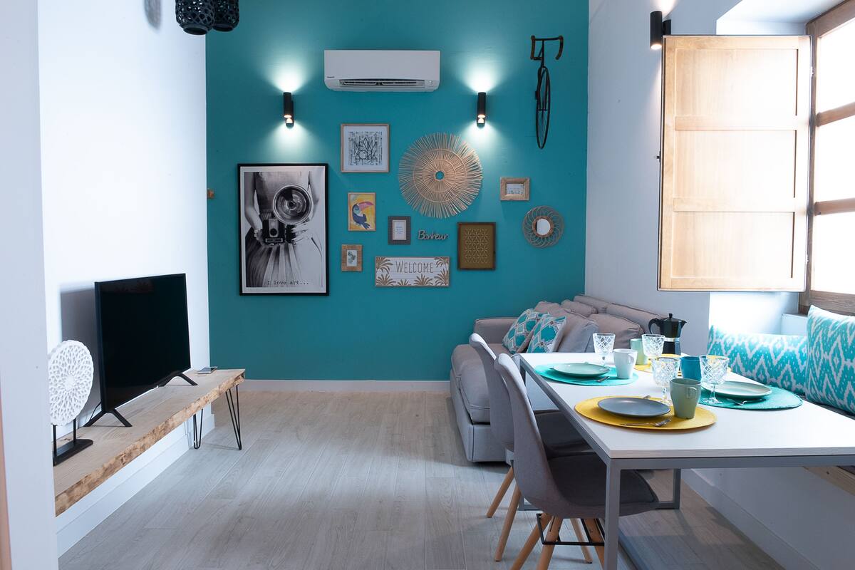 Un salon moderne avec des murs de couleur turquoise, un canapé, une table à manger dressée avec des assiettes et des verres, et un climatiseur fixé au mur.