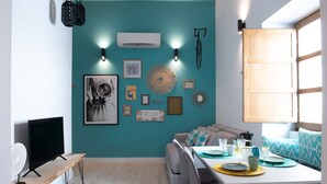 En moderne stue med teal farvede vægge, en sofa, et spisebord dækket med tallerkener og glas, samt diverse vægdekorationer, heriblandt en cykel og et velkommenskilt.