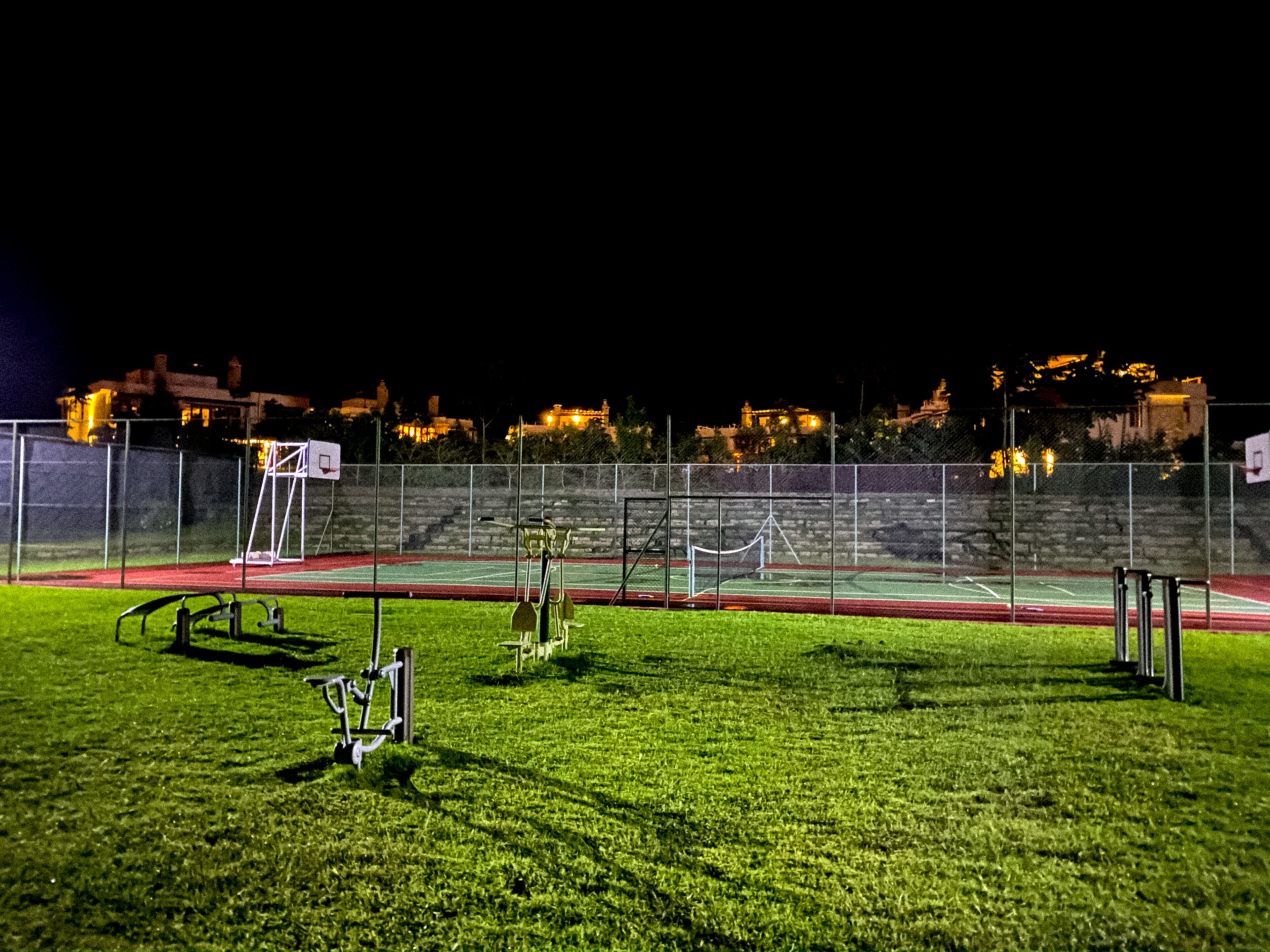 Een sportfaciliteit buiten, 's nachts, met een basketbalveld, trainingsapparatuur en een hek.