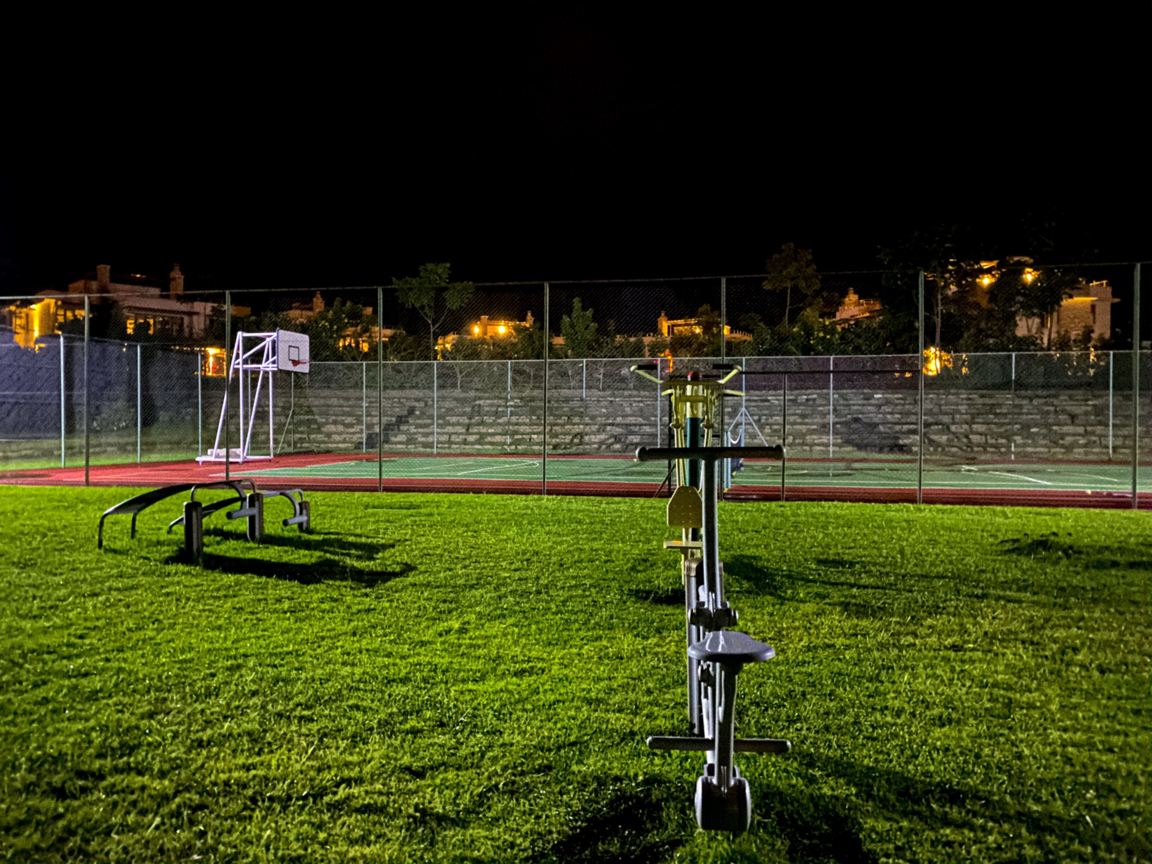 Un terrain de basket-ball en plein air, éclairé la nuit, avec une clôture, un panier de basket et du matériel d'exercice.