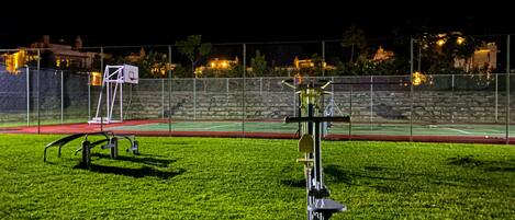 Un terrain de basket-ball en plein air, éclairé la nuit, avec une clôture, un panier de basket et du matériel d'exercice.