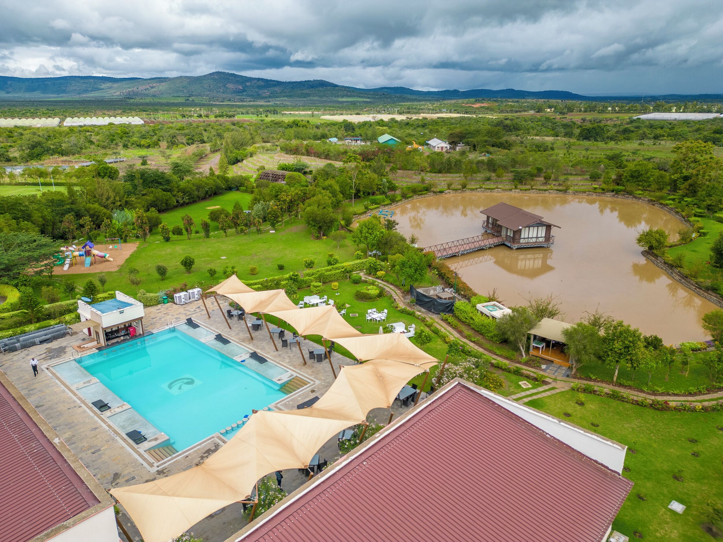 A large swimming pool with sun umbrellas, a small building near a pond, and a lush green landscape with distant hills.