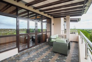 Un balcón con vista al paisaje, una zona de estar con sofá y sillones, y un ventanal de vidrio que da al interior.