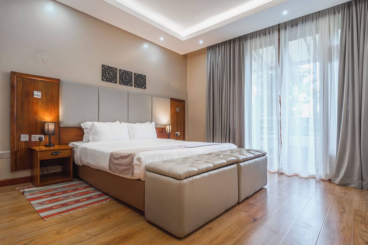 Un dormitorio moderno con una cama grande, una mesita de noche de madera, un banco y un amplio ventanal con cortinas.