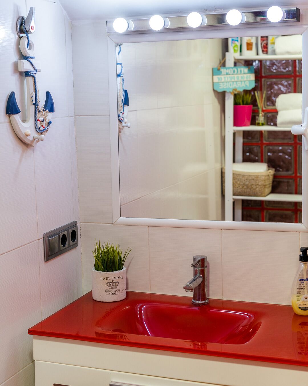 A bathroom with a red sink, a mirror with lights, a towel rack, and a small plant.