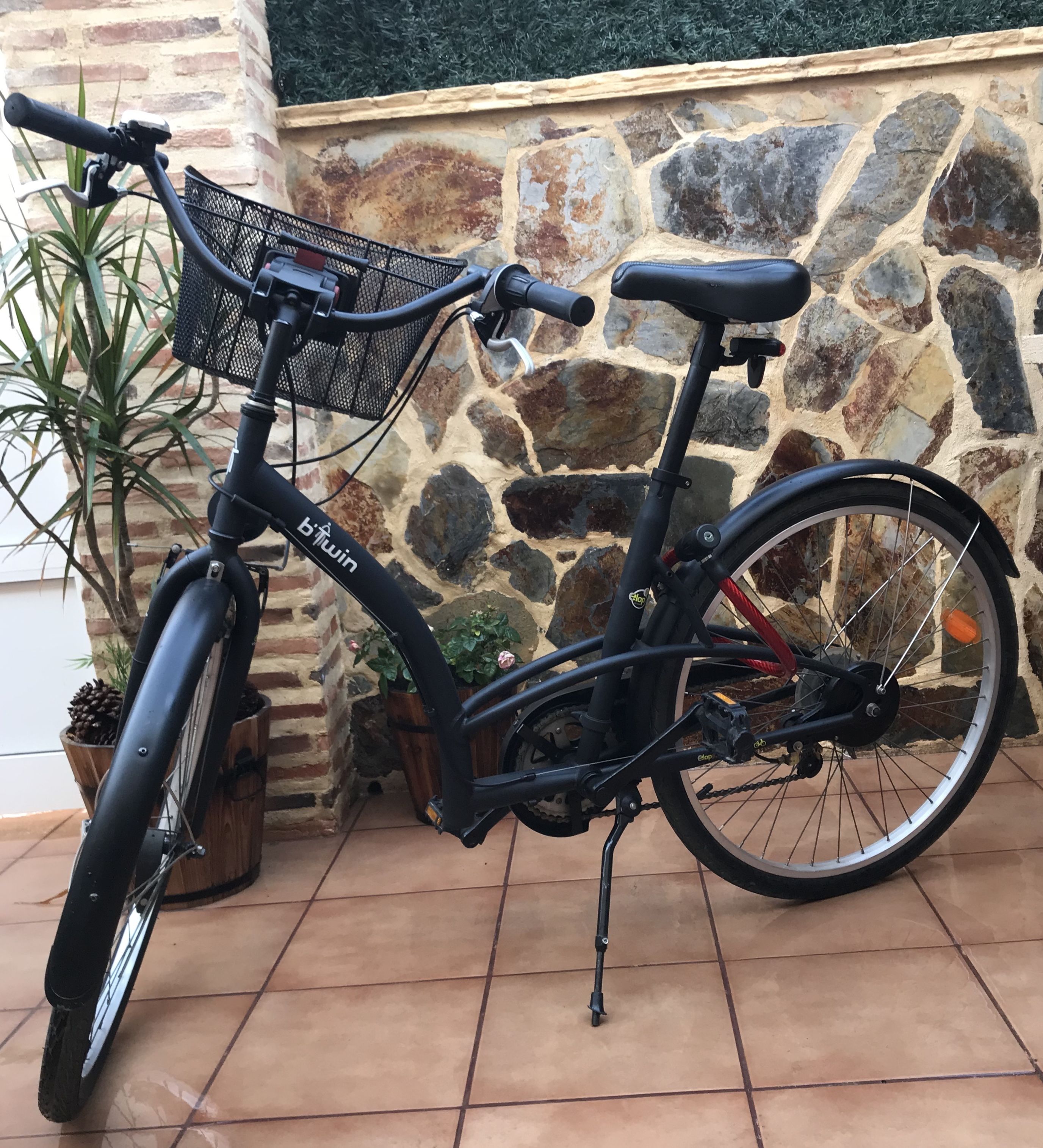 Una bicicleta estática con una canasta y portabebidas para agua, estacionada junto a una pared de piedra y una planta en maceta cerca.