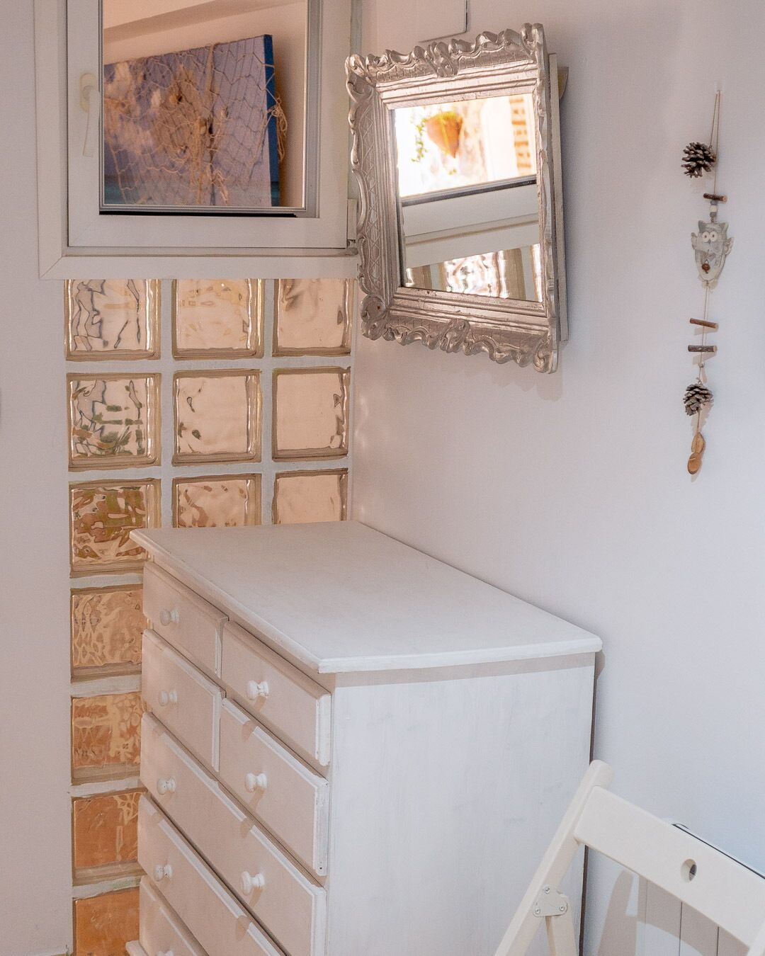 Un tocador blanco con cajones frente a una pared con un espejo decorativo y un pequeño estante con un adorno de piña.