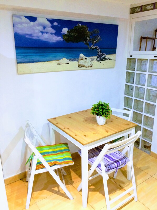 A small dining area with a wooden table and two white chairs, a potted plant on the table, and a beach-themed painting on the wall.