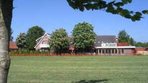 A building with a red roof and white walls, surrounded by a grassy field and trees. - Landgasthaus Fecht (Aurich)