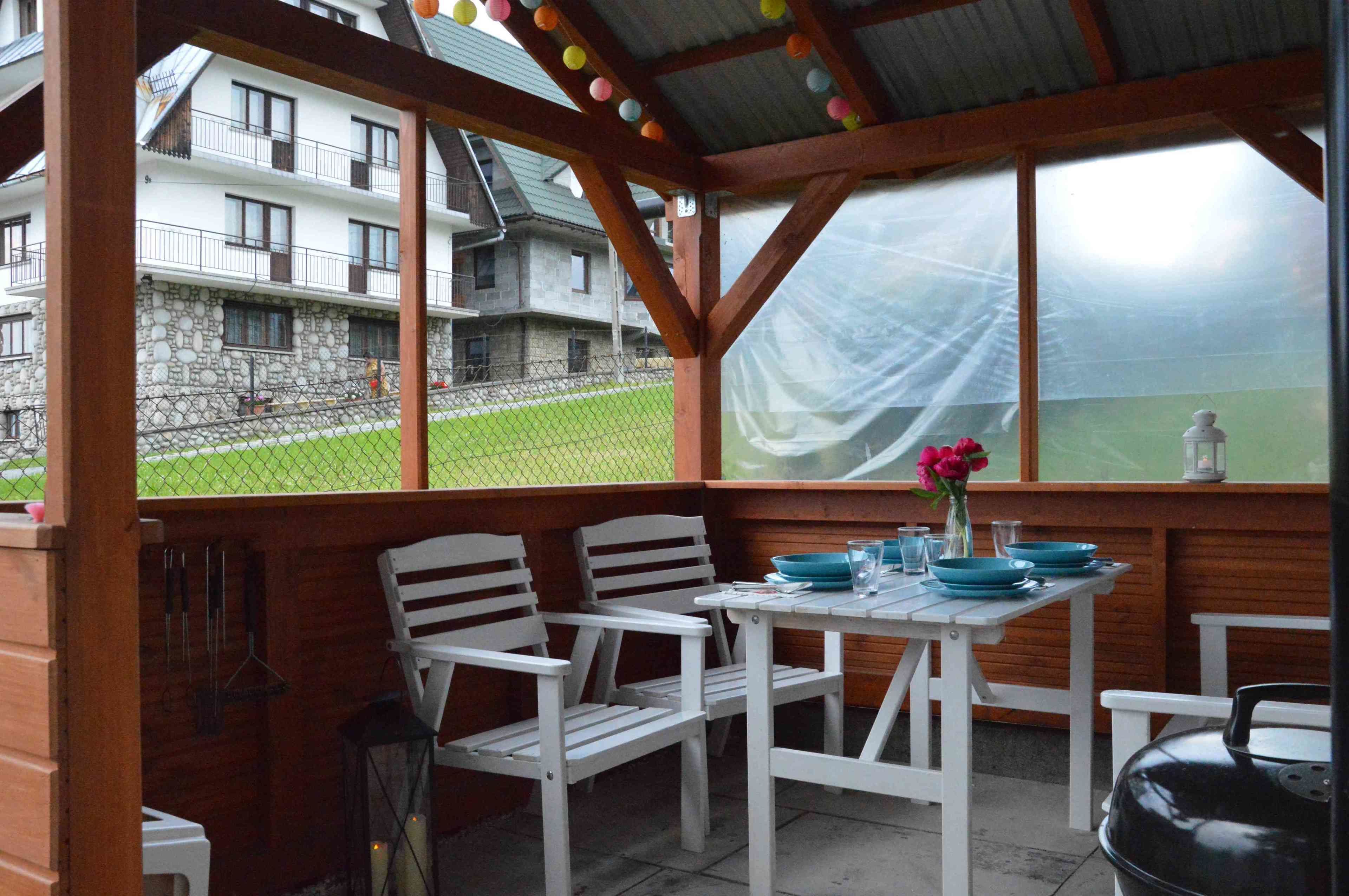 An outdoor patio with white chairs and a table, a covered area with wooden beams, and a view of a building with a stone facade.
