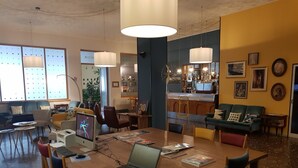 A modern meeting room with a wooden table, laptops, and chairs. There are large windows with blinds, a bar area, and a green sofa. - ibis styles Trani (Trani)