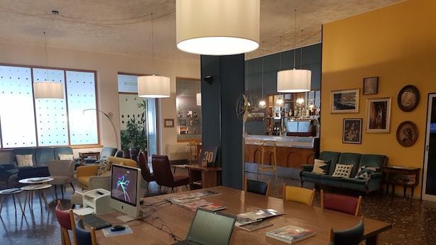 A modern meeting room with a wooden table, laptops, and chairs. There are large windows with blinds, a bar area, and a green sofa.