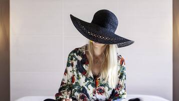 Una persona seduta su un letto con un libro, indossa una camicia floreale e un cappello grande.