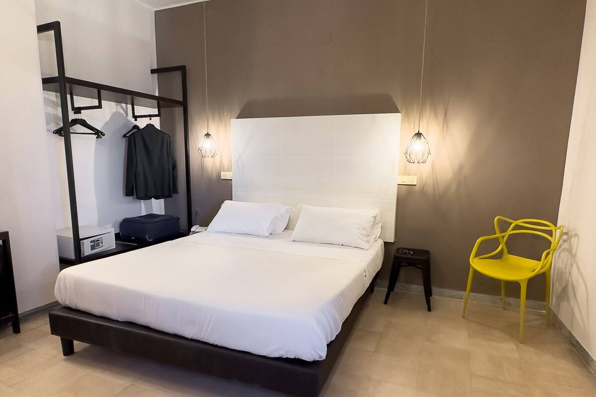 A modern bedroom with a large bed, a bedside table, a mirror, a hanging rack, and a yellow chair.