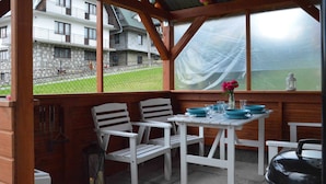 An outdoor patio with white chairs and a table, a covered area with wooden beams, and a view of a building with a stone facade. - Maryna House - Widokowy Apartament (Zakopane)