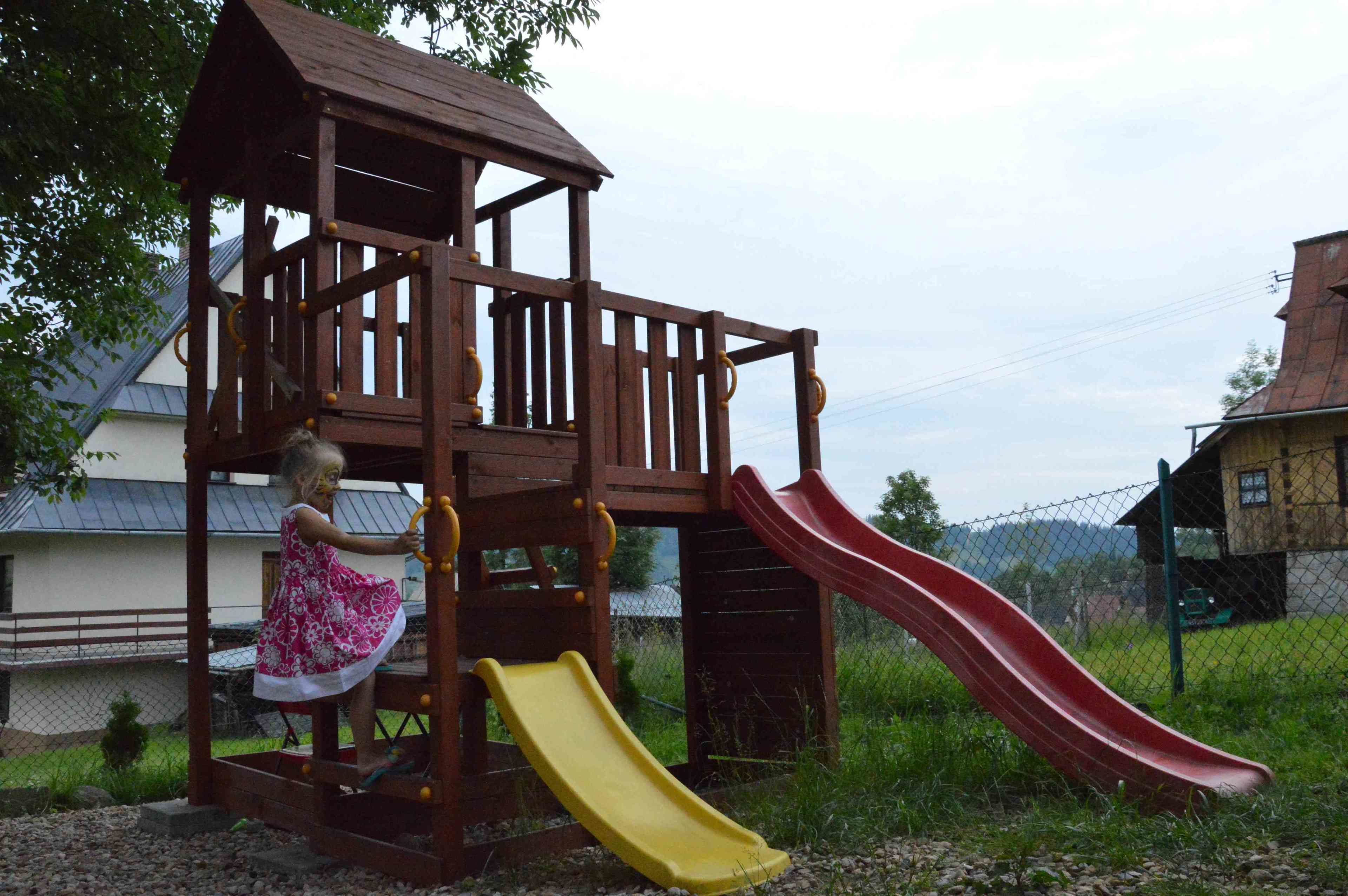A wooden play structure with a red and yellow slide, surrounded by a fence and grassy area.