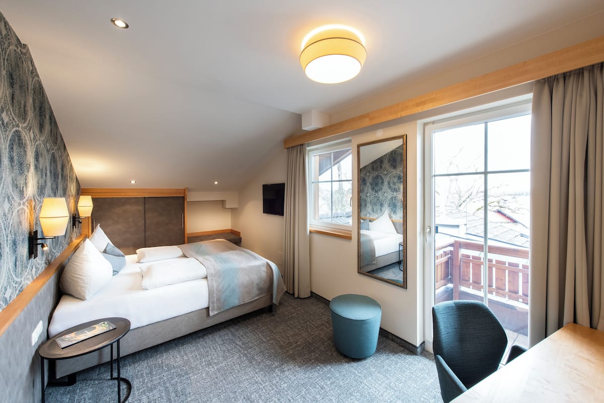 Habitación de hotel con cama, una mesita, una silla, un reposapiés redondo, un ventanal con cortinas, un espejo y una lámpara fijada a la pared.