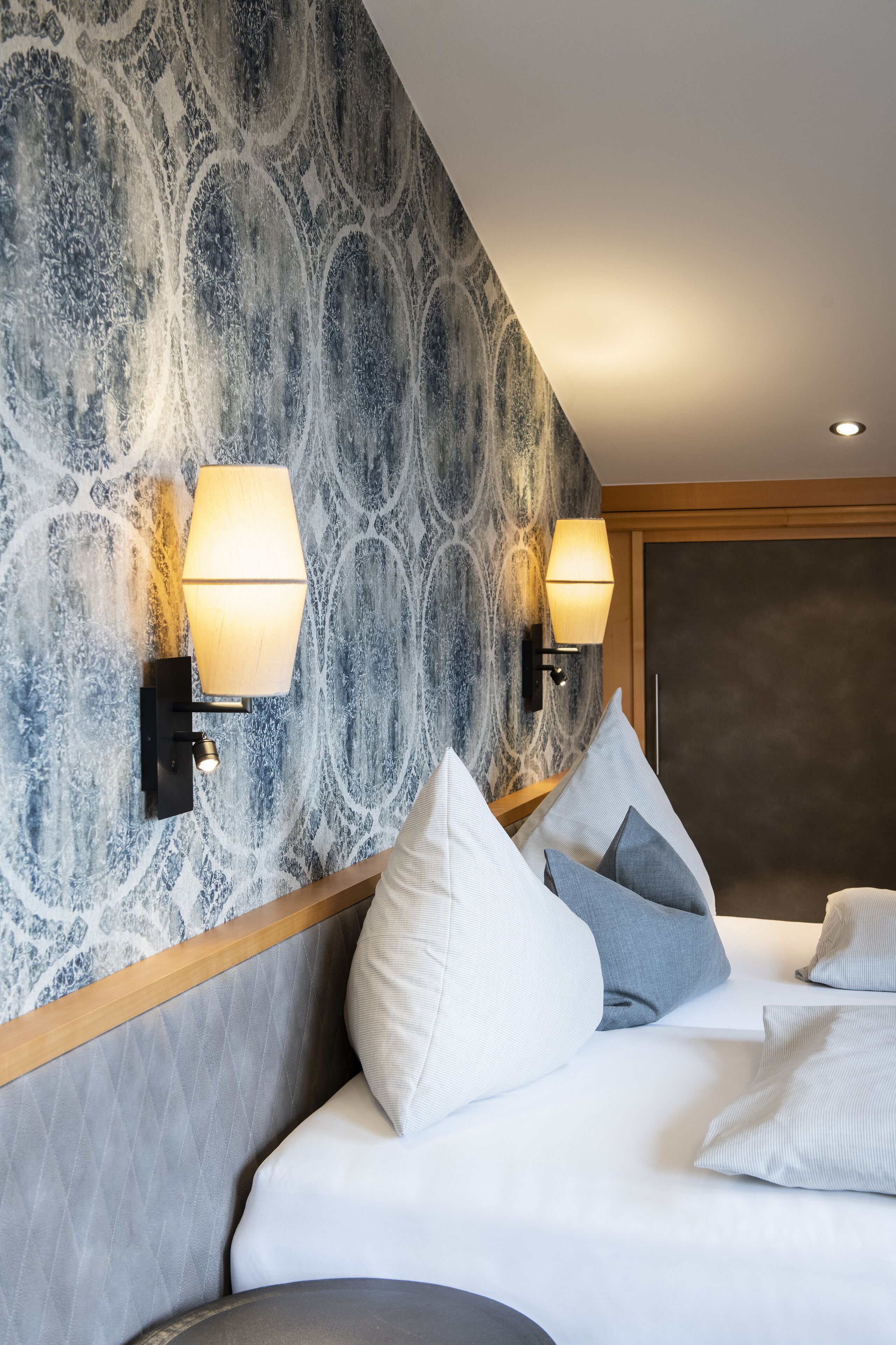 A hotel room with a patterned wall, two wall-mounted lamps, a bed with pillows, and a wooden headboard.