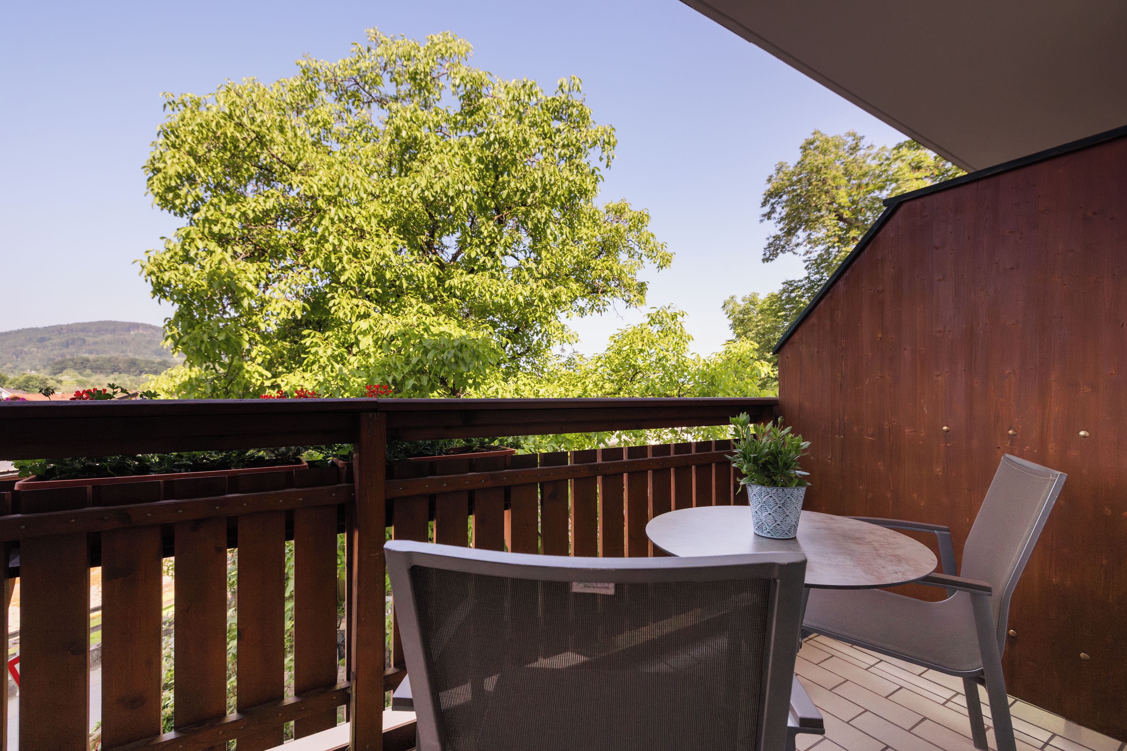 A wooden deck with a table and chairs, overlooking a large tree and a hilly landscape.