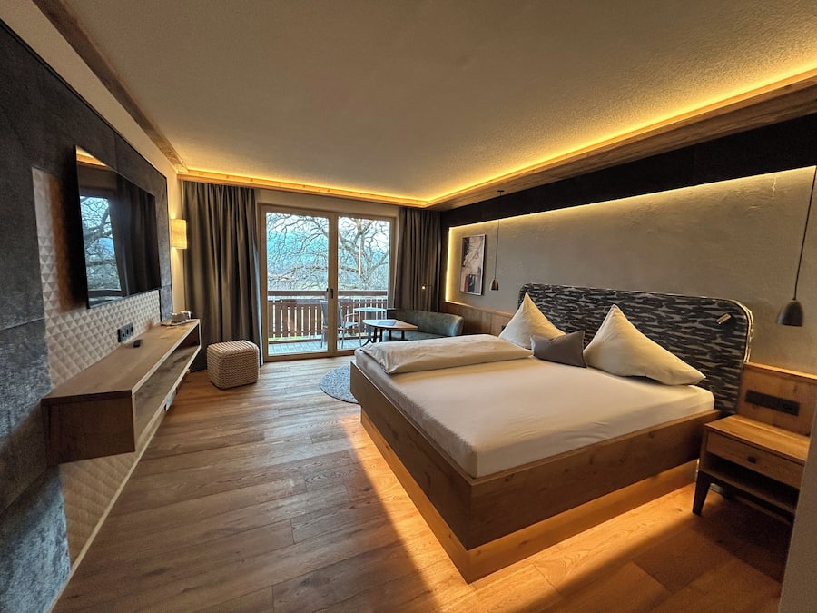 Una habitación de hotel moderna con piso de madera, una cama grande con dos almohadas, un escritorio y una silla. Hay una puerta corrediza de vidrio que da a un espacio exterior con terraza de madera.