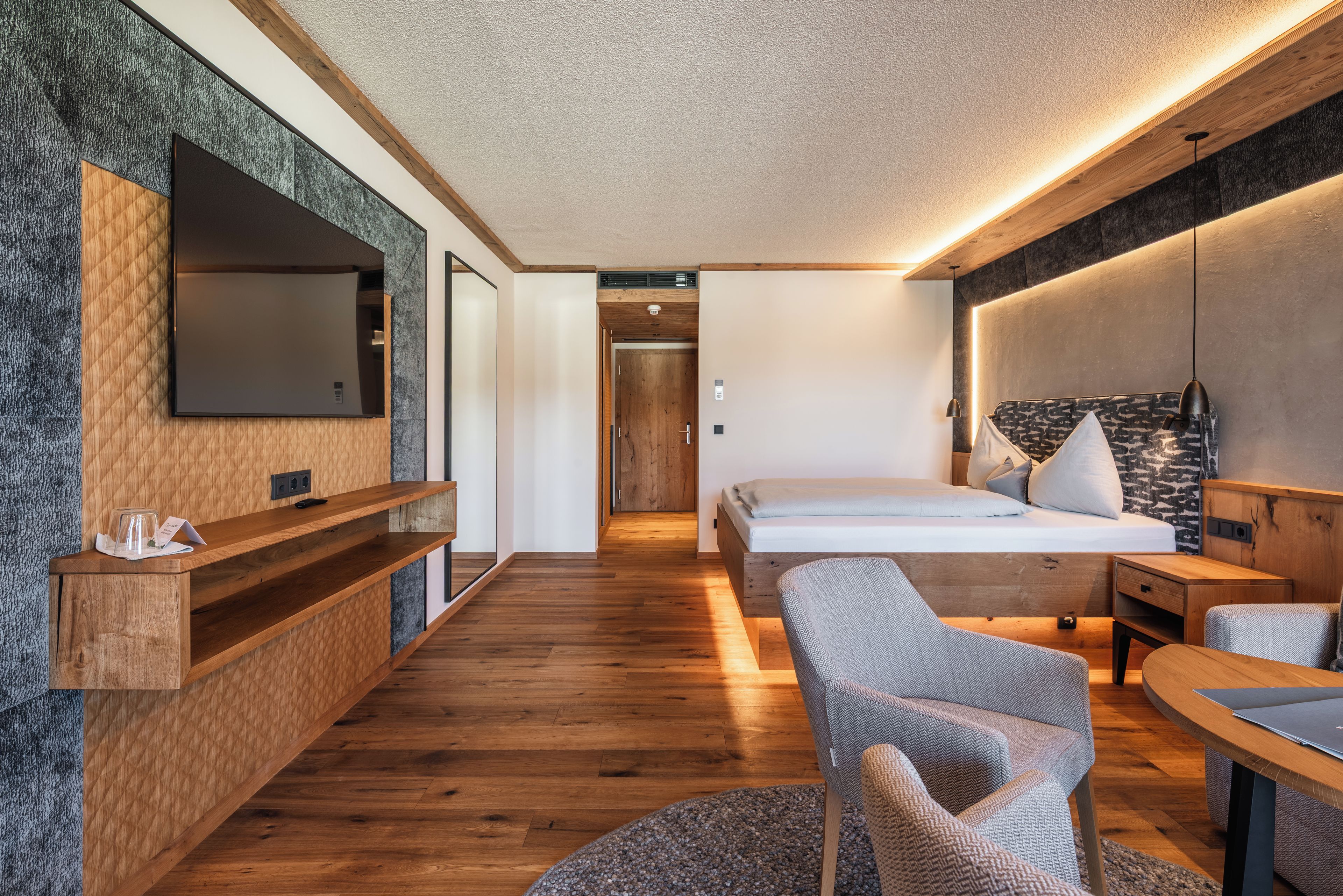 A modern hotel room with a large bed, a desk, a chair, a TV, and a wooden floor.