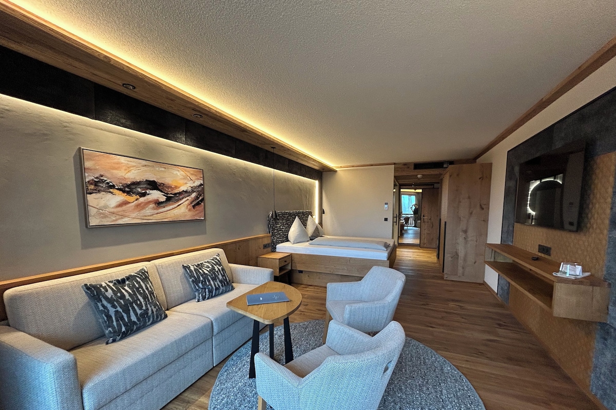 A modern hotel room with a sofa, two armchairs, a small table, a bed, and a TV.