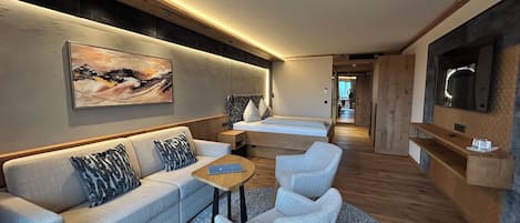 A modern hotel room with a sofa, two armchairs, a small table, a bed, and a TV.