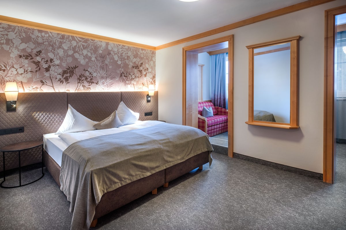 Habitación de hotel con cama, mesita de noche, lámpara, papel tapiz con motivos florales, espejo y vista a otra habitación.