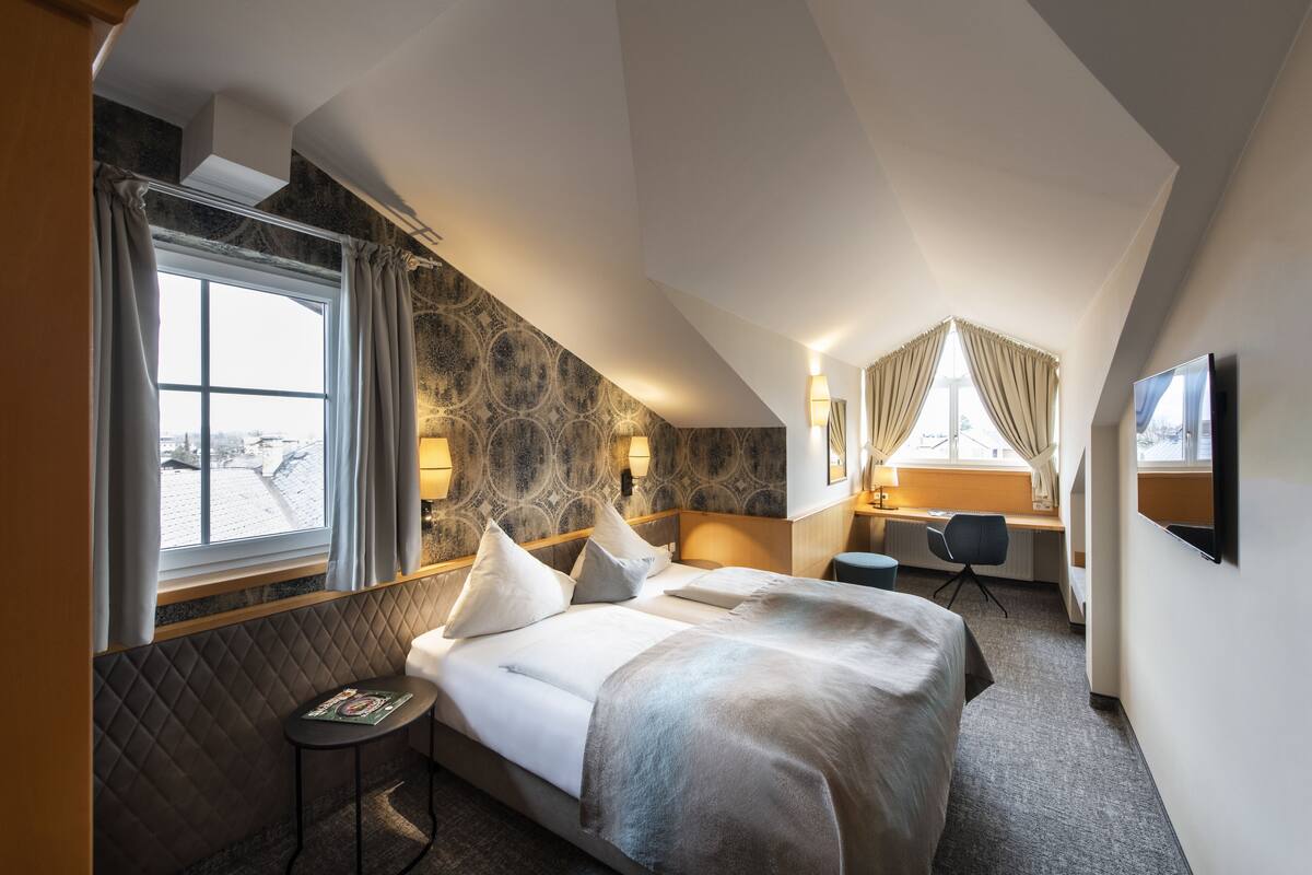 Habitación de hotel con una cama grande, un escritorio y una silla. Se ve una ventana con cortinas, un televisor montado en la pared y una pequeña mesa con una revista.