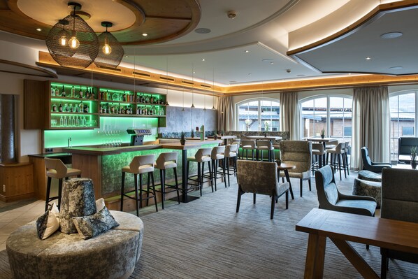 Zona de bar con mostrador iluminado en verde, diversas opciones de asientos, incluyendo taburetes y sillas, y vistas a través de amplios ventanales.