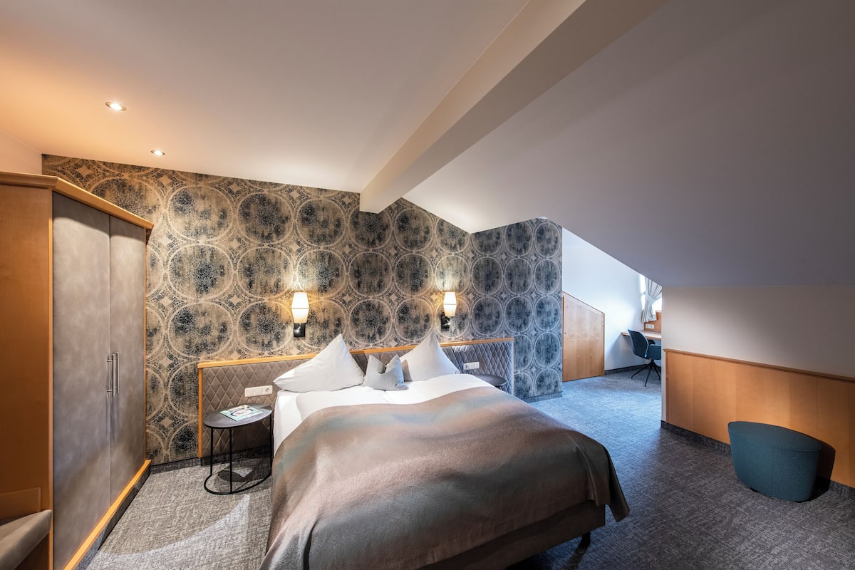 Una habitación de hotel moderna con una cama grande, una mesita pequeña y una silla. La habitación tiene un papel tapiz estampado y un cabecero de madera.